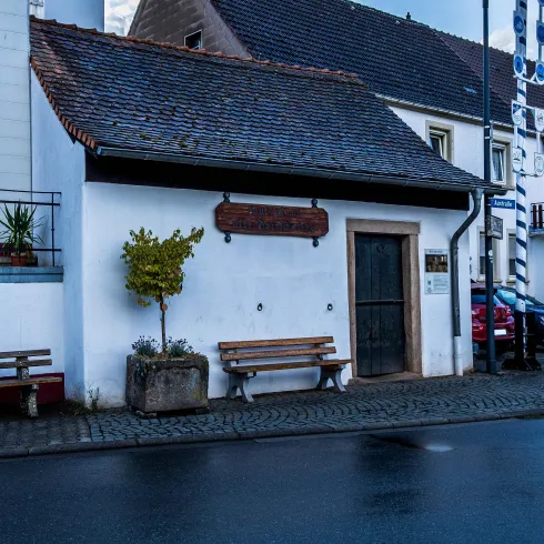 Ein kleines, charmantes Haus mit einem dunklen Dach und einer weißen Fassade. Vor dem Haus stehen zwei Bänke und ein kleiner Baum.