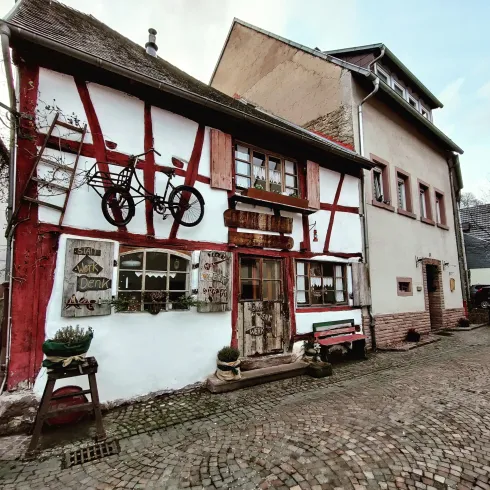Ein charmantes Fachwerkhaus mit roter Fachwerkkonstruktion und Fensterläden. Auf dem Gehweg steht eine Bank und an der Fassade hängt ein Fahrrad.