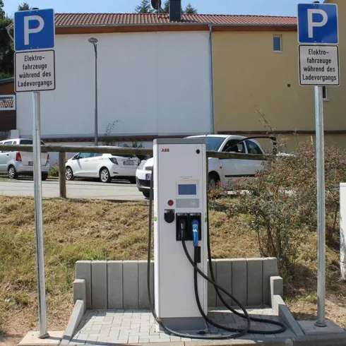 Eine E-Ladestation für Elektrofahrzeuge mit zwei Parkplätzen. Im Hintergrund sind einige Autos und Häuser zu sehen.