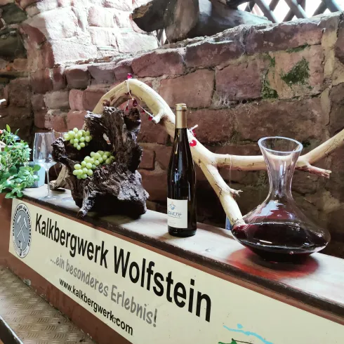 Eine rustikale Tischdekoration mit einem Weinflasche, einer Karaffe und dekorativen Elementen wie Weintrauben und Holz. Im Hintergrund ist ein Schild mit dem Namen Kalkbergwerk Wolfstein zu sehen.