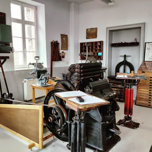 Ein Raum mit historischen Druckmaschinen und Schreibgeräten. Die Wände sind mit verschiedenen Druckmaterialien und Werkzeugen dekoriert.