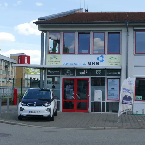 Ein modernes Mobilitätszentrum mit einem Elektroauto vor dem Eingang. Die Fassade ist hell und freundlich gestaltet.