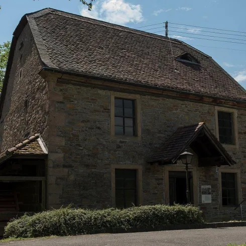 Ein altes, steinernes Gebäude mit einem Satteldach und Fensterläden. Im Vordergrund sind grüne Sträucher und ein Weg zu sehen.