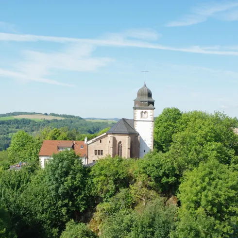 Eine Kirche auf einem Hügel, umgeben von grünen Bäumen. Der Himmel ist klar und blau.