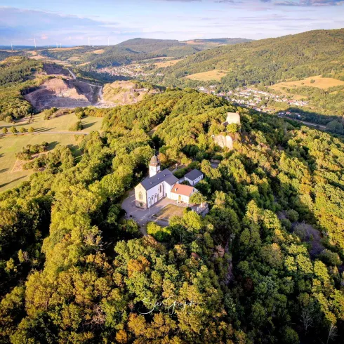 Eine malerische Landschaft mit Wäldern und sanften Hügeln. Auf dem Hügel steht eine alte Kirche, umgeben von grüner Natur.
