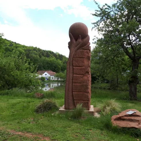 Eine große Skulptur aus rotem Stein, die eine Hand mit einer Kugel darstellt. Im Hintergrund sind Bäume und ein Teich zu sehen.