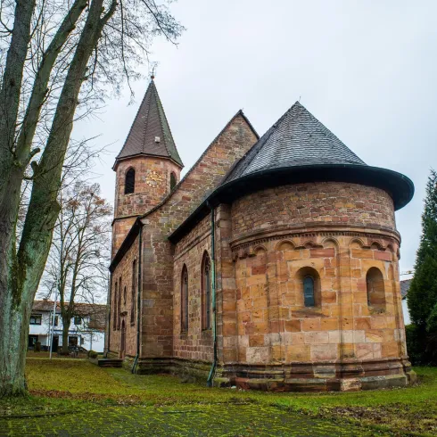 Eine alte Kirche mit einem schmalen Turm und schönem Mauerwerk aus rotem Sandstein. Umgeben von Bäumen und einem ruhigen Garten.