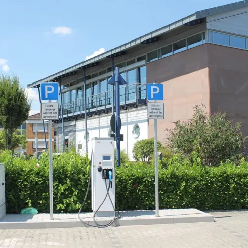 Eine E-Ladesäule steht auf einem Parkplatz vor einem modernen Gebäude. Im Hintergrund sind Bäume und ein blauer Himmel sichtbar.