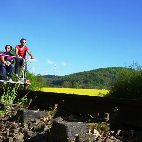 Eine Gruppe von vier Personen fährt auf einem ungewöhnlichen Fahrzeug entlang von Schienen. Im Hintergrund ist eine grüne Landschaft und ein klarer blauer Himmel zu sehen.