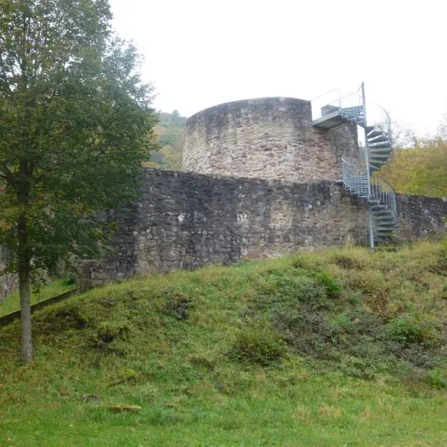 Eine alte Steinmauer mit einem runden Turm und einer Stahlspiraltreppe. Die umgebende Landschaft ist grün und bewaldet.