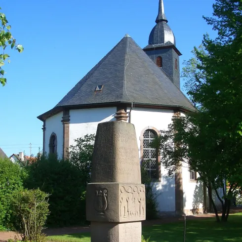 Eine kleine Kirche mit einem spitzen Turm und einem historischen Stein im Vordergrund. Der Garten ist grün und einladend.